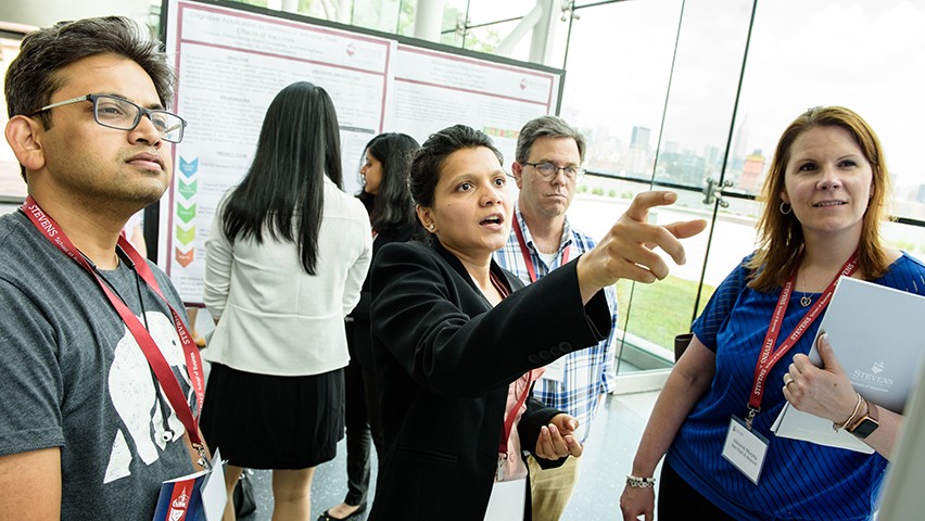 A group of students and professionals looking at a poster presentation at Stevens, with the New York City skyline in the background.