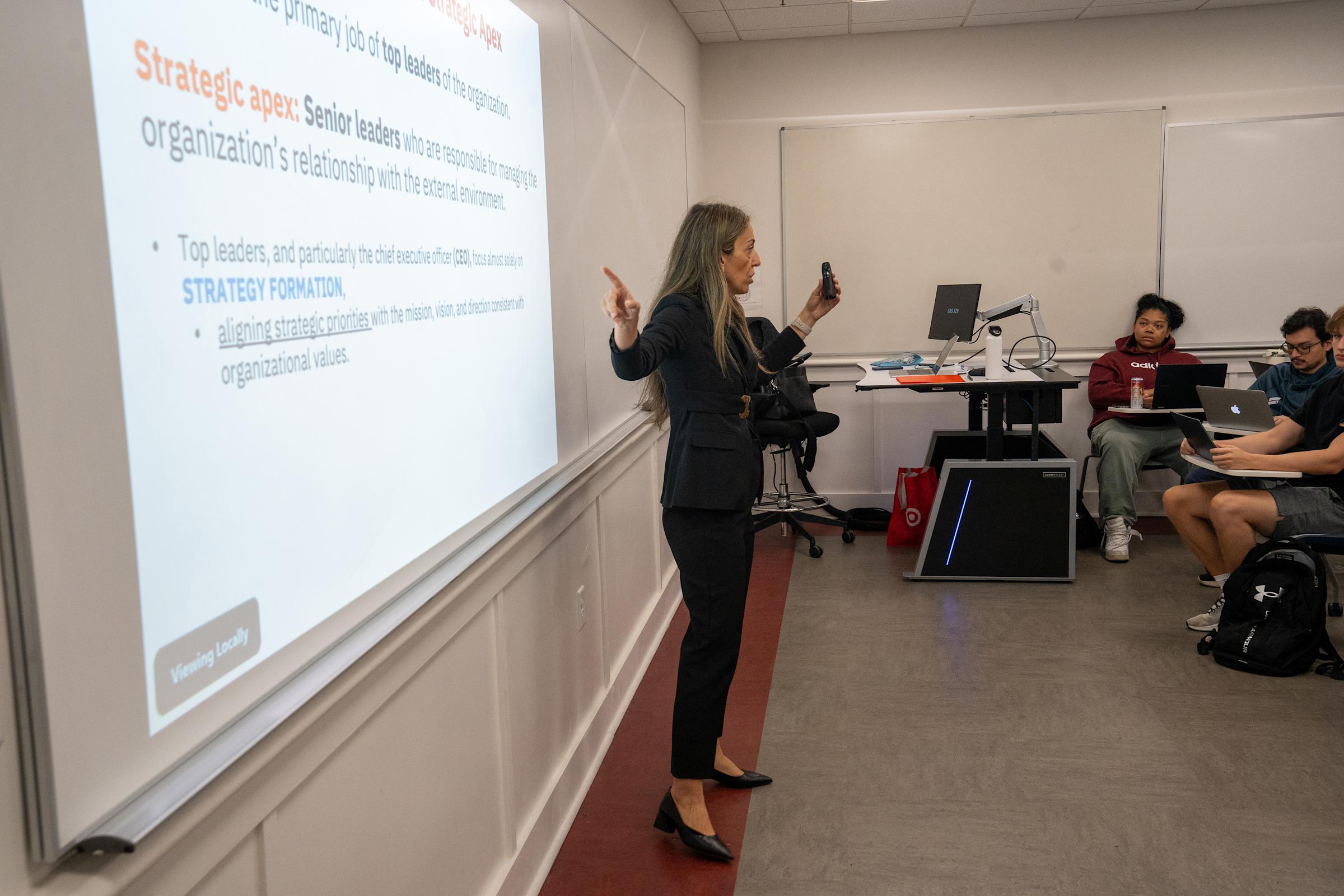 A woman in a dark business suit presents to students in a classroom, gesturing toward a projected slide about strategic leadership and organizational strategy formation. Students with laptops sit at tiered desks listening to the lecture.