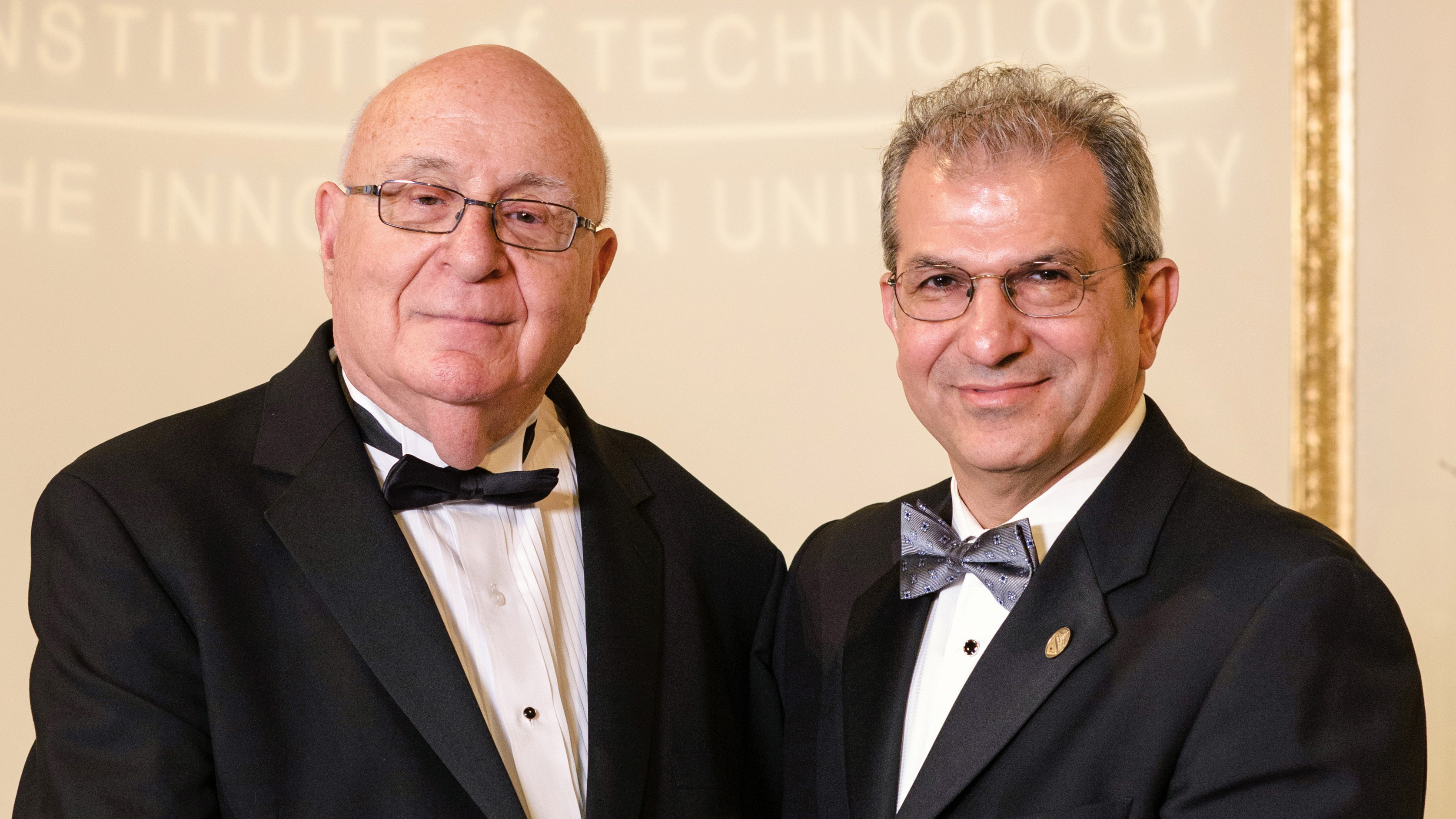 Photo of Dave Farber ’56 M.S. ’61, left, being presented a plaque by Stevens President Nariman Farvardin on a stage, with the Stevens logo and name projected in the background.