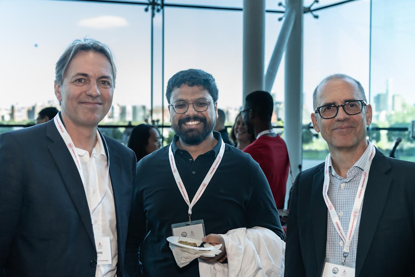 Three men pose together at a conference networking event in a modern glass-walled venue.