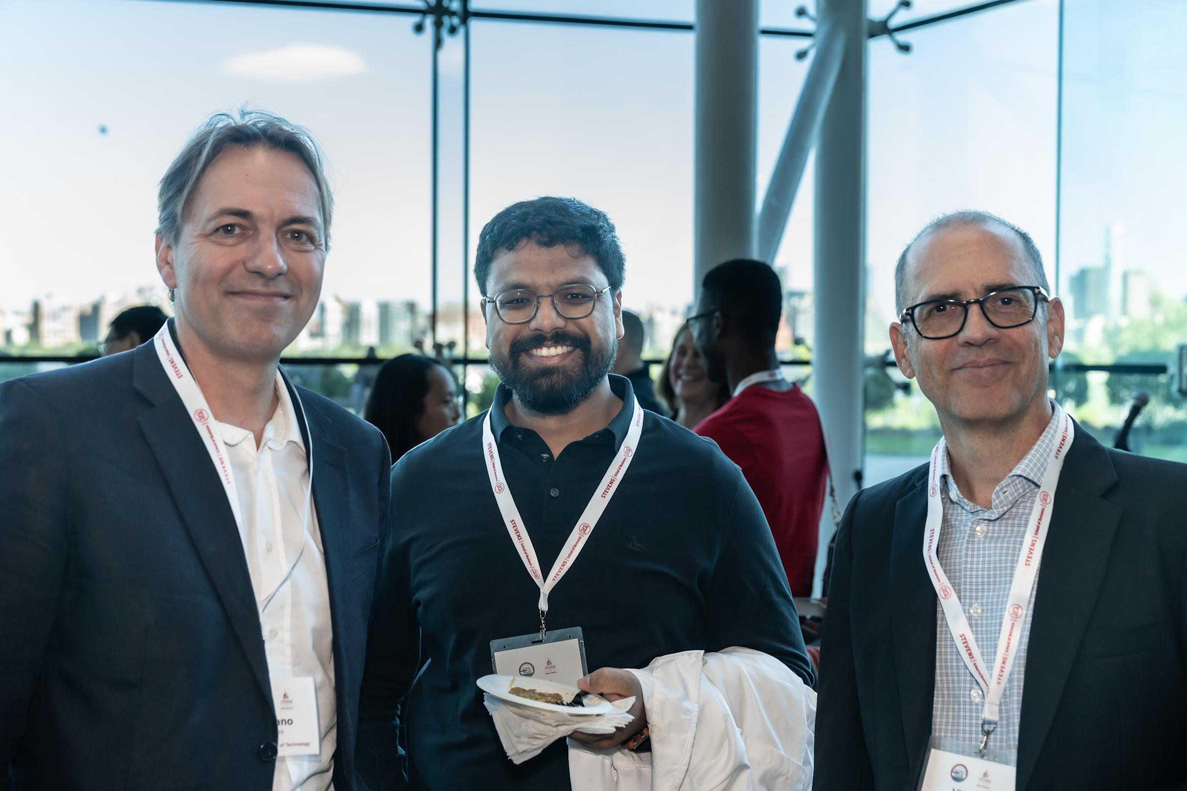 Three men pose together at a conference networking event in a modern glass-walled venue.