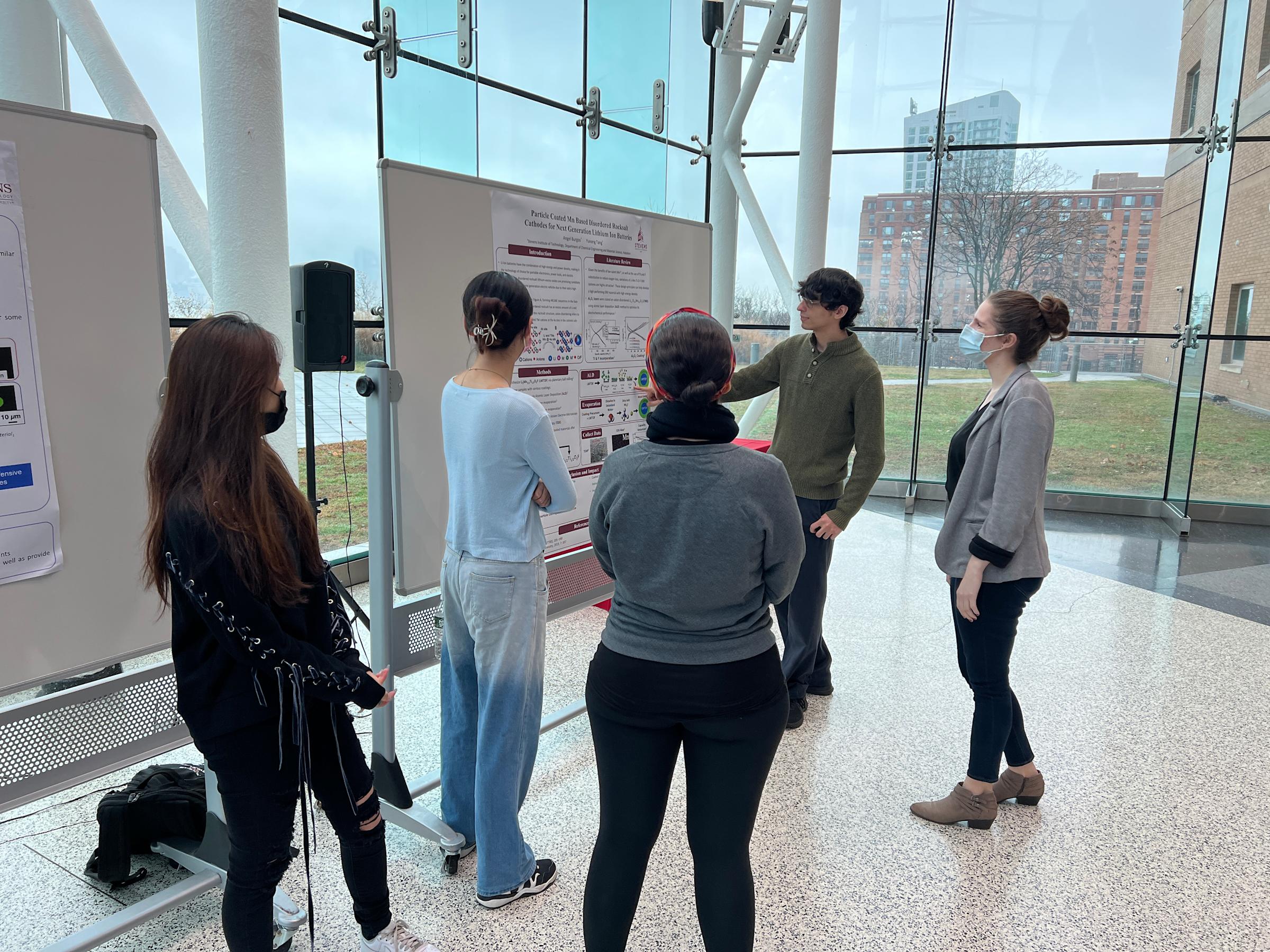 Angel Burgos presents his poster to other students in front of huge window overlooking Manhattan