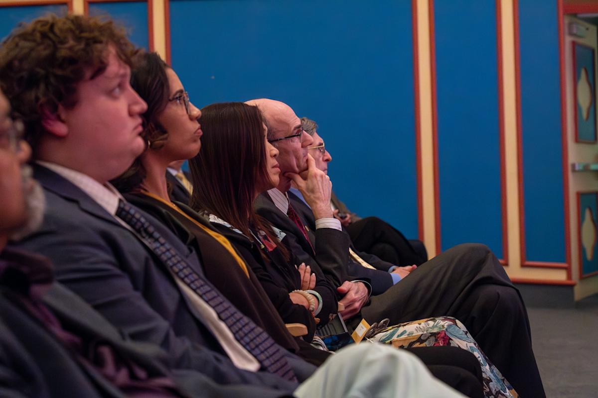 Six people sit in the audience listening, wearing business clothes.