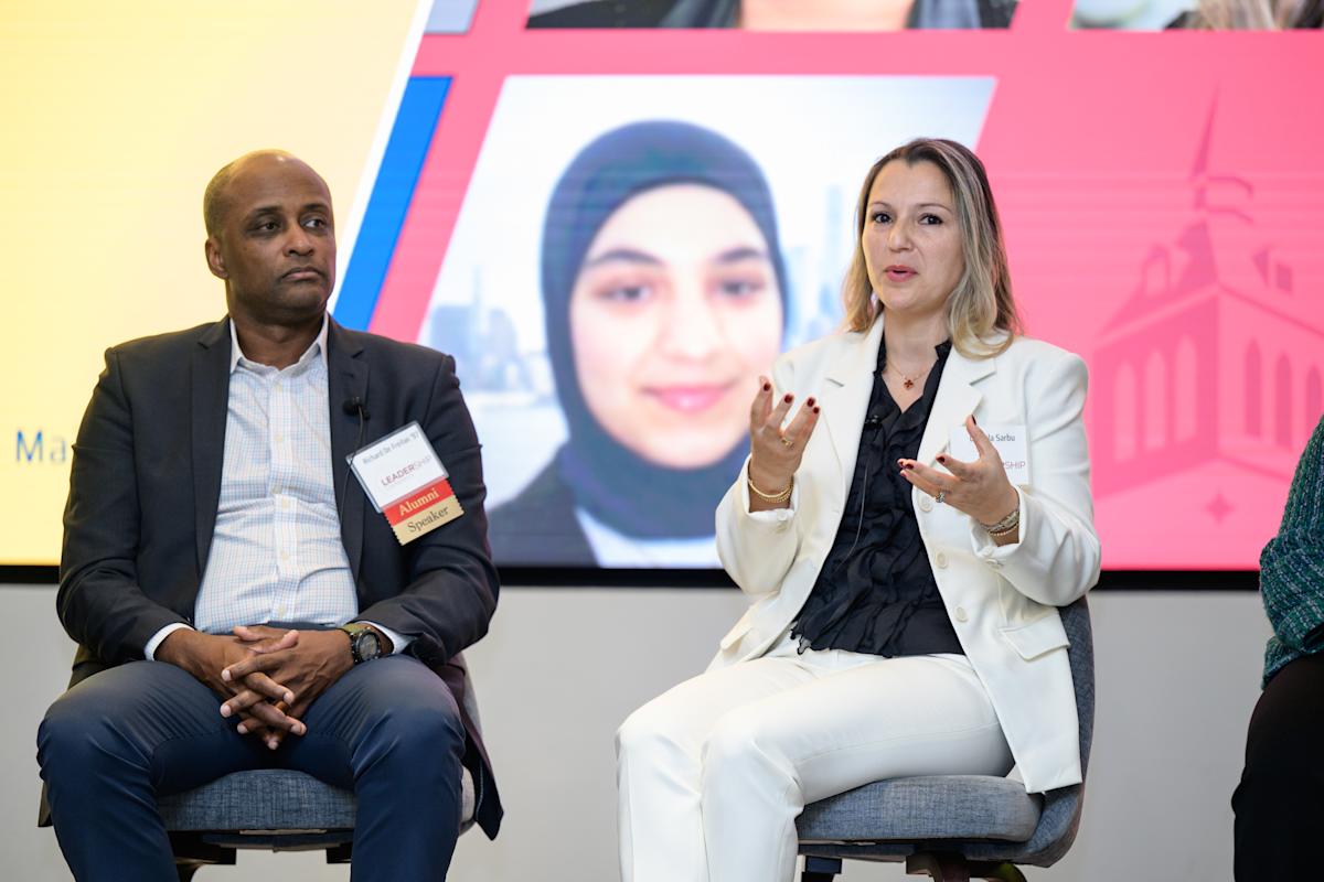 Two panelists sit on stage during a professional event. One speaker in a light-colored suit gestures while speaking, while another panelist listens. A large screen behind them displays event graphics.