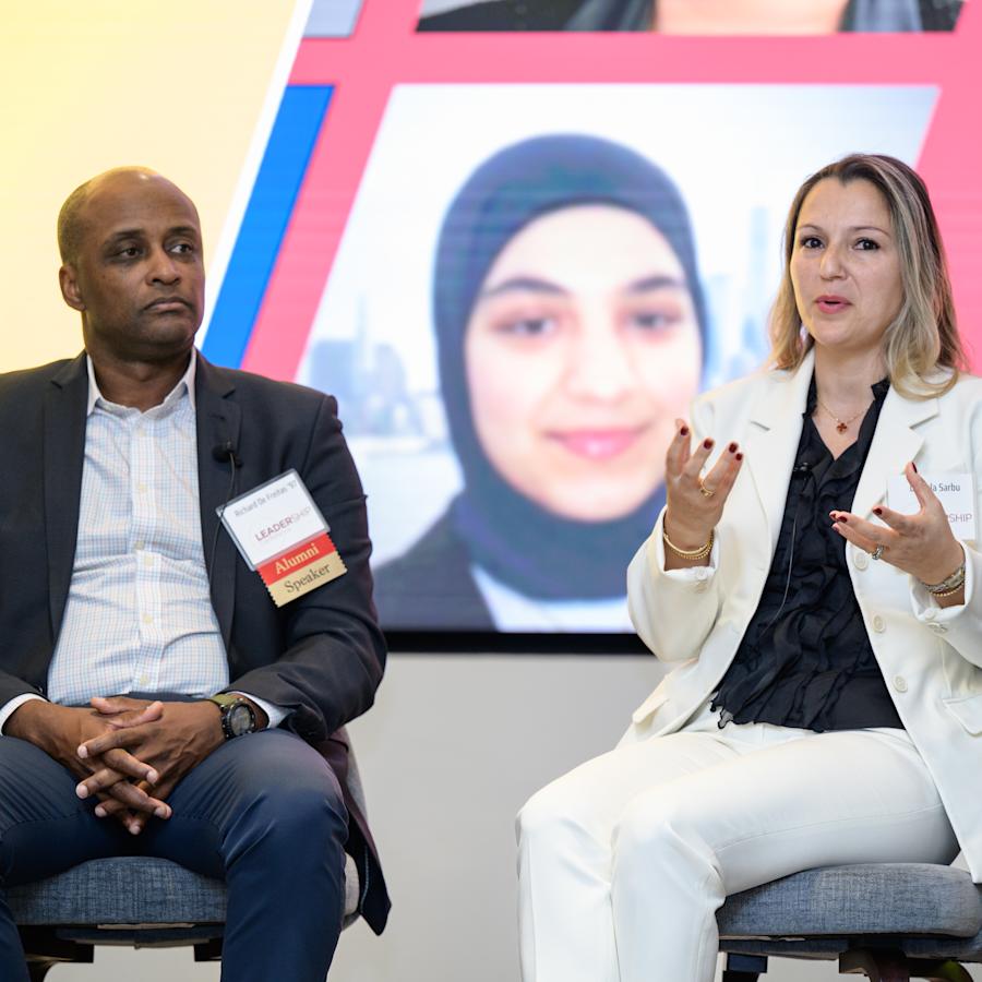 Two panelists sit on stage during a professional event. One speaker in a light-colored suit gestures while speaking, while another panelist listens. A large screen behind them displays event graphics.