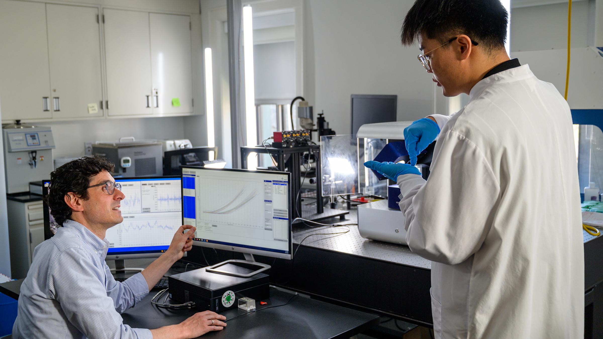 Professor in lab gestures to information on monitor while student looks on.