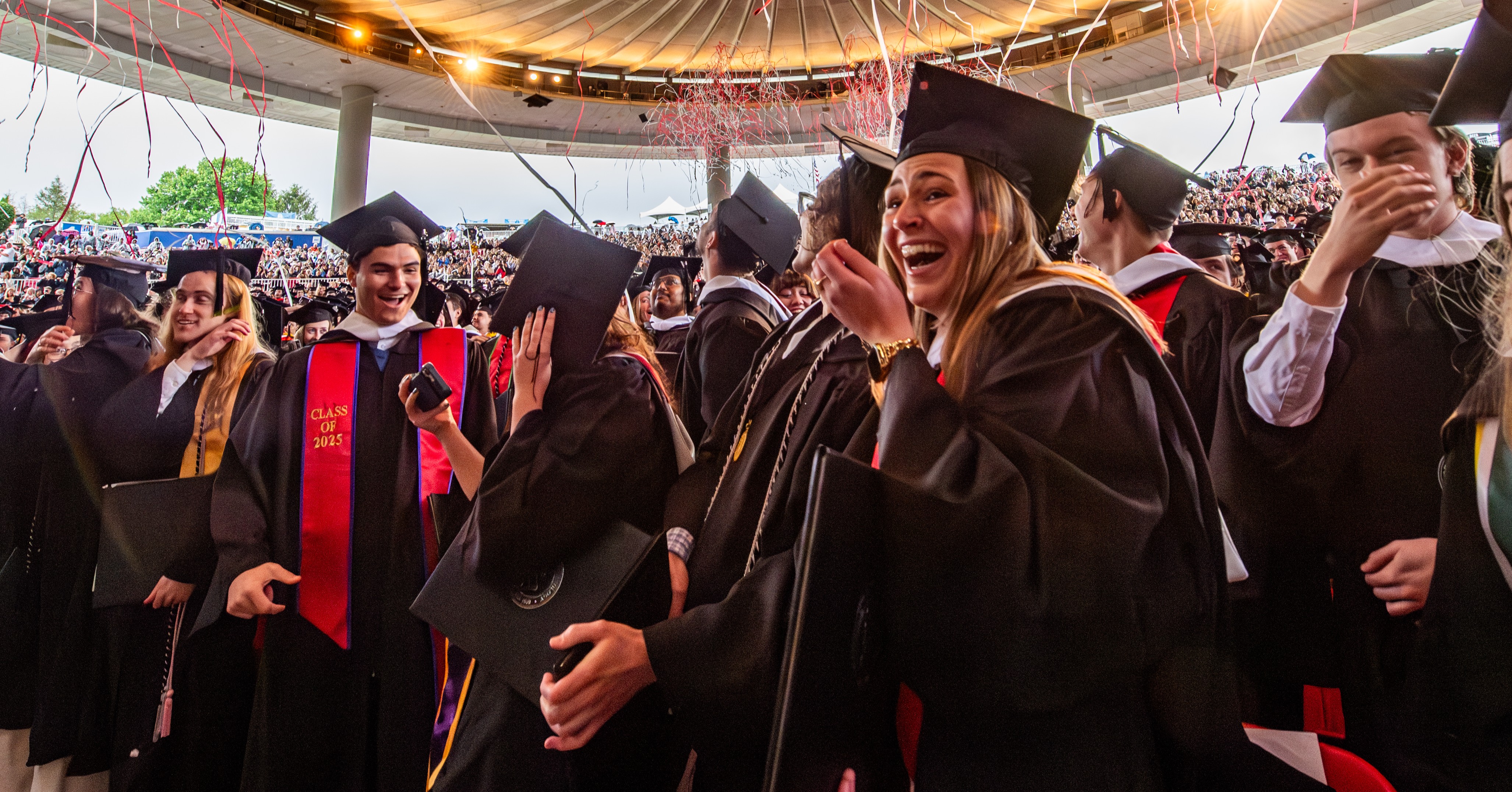 A large group of students inside a amphitheater wear gap and gowns and celebrate graduation. There are streams in the air.