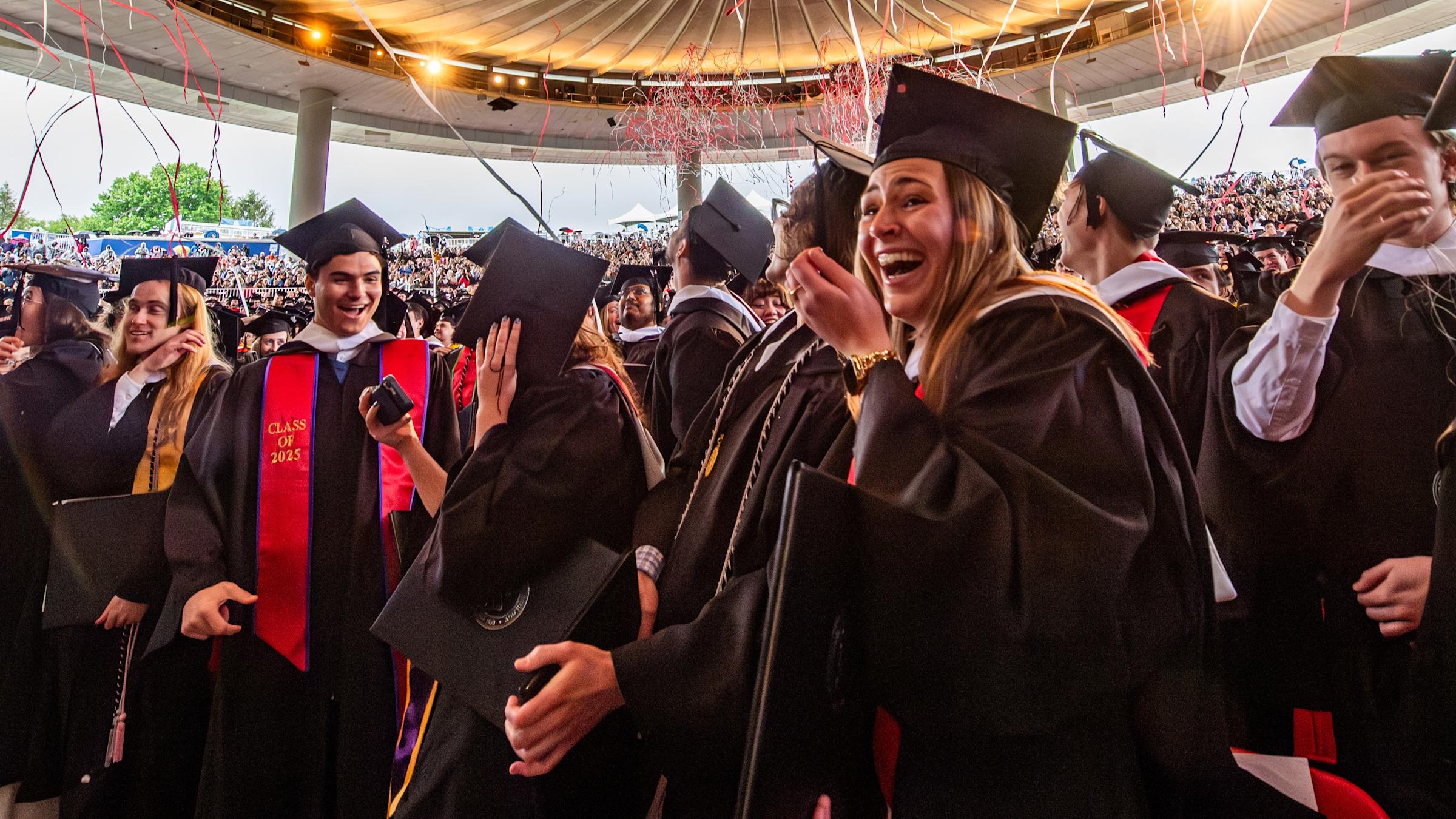 A large group of students inside a amphitheater wear gap and gowns and celebrate graduation. There are streams in the air.