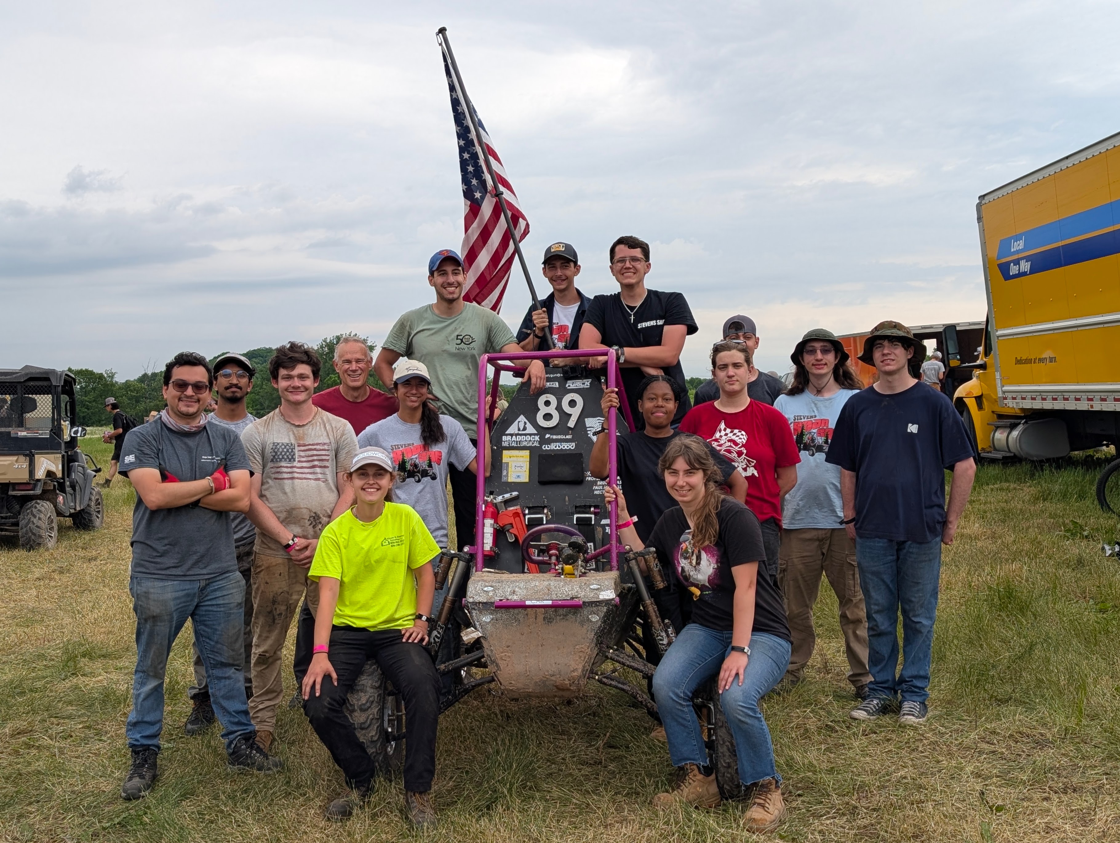 Members of Stevens Institute of Technology’s Society of Automotive Engineers pose with their racing vehicle in an open field, gathered around the student-built off-road car beneath an American flag during a competition event.