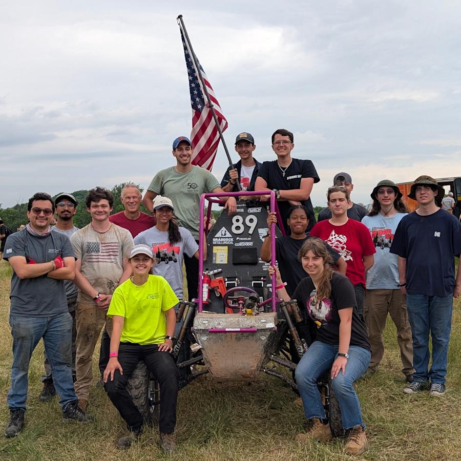 Members of Stevens Institute of Technology’s Society of Automotive Engineers pose with their racing vehicle in an open field, gathered around the student-built off-road car beneath an American flag during a competition event.