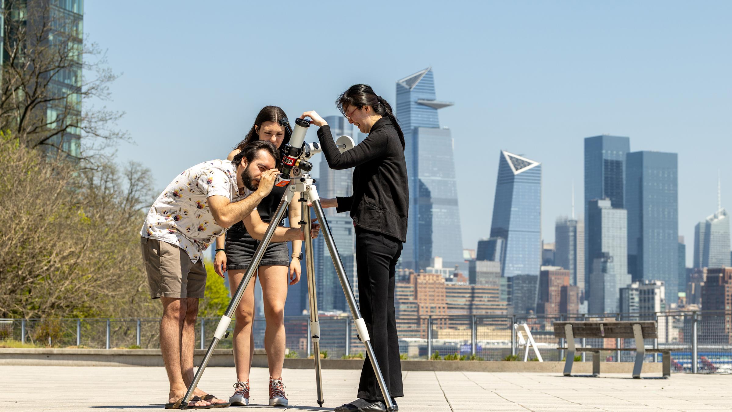 Two students and a professor work with a telescope on campus with the Manhattan skyline in the background.