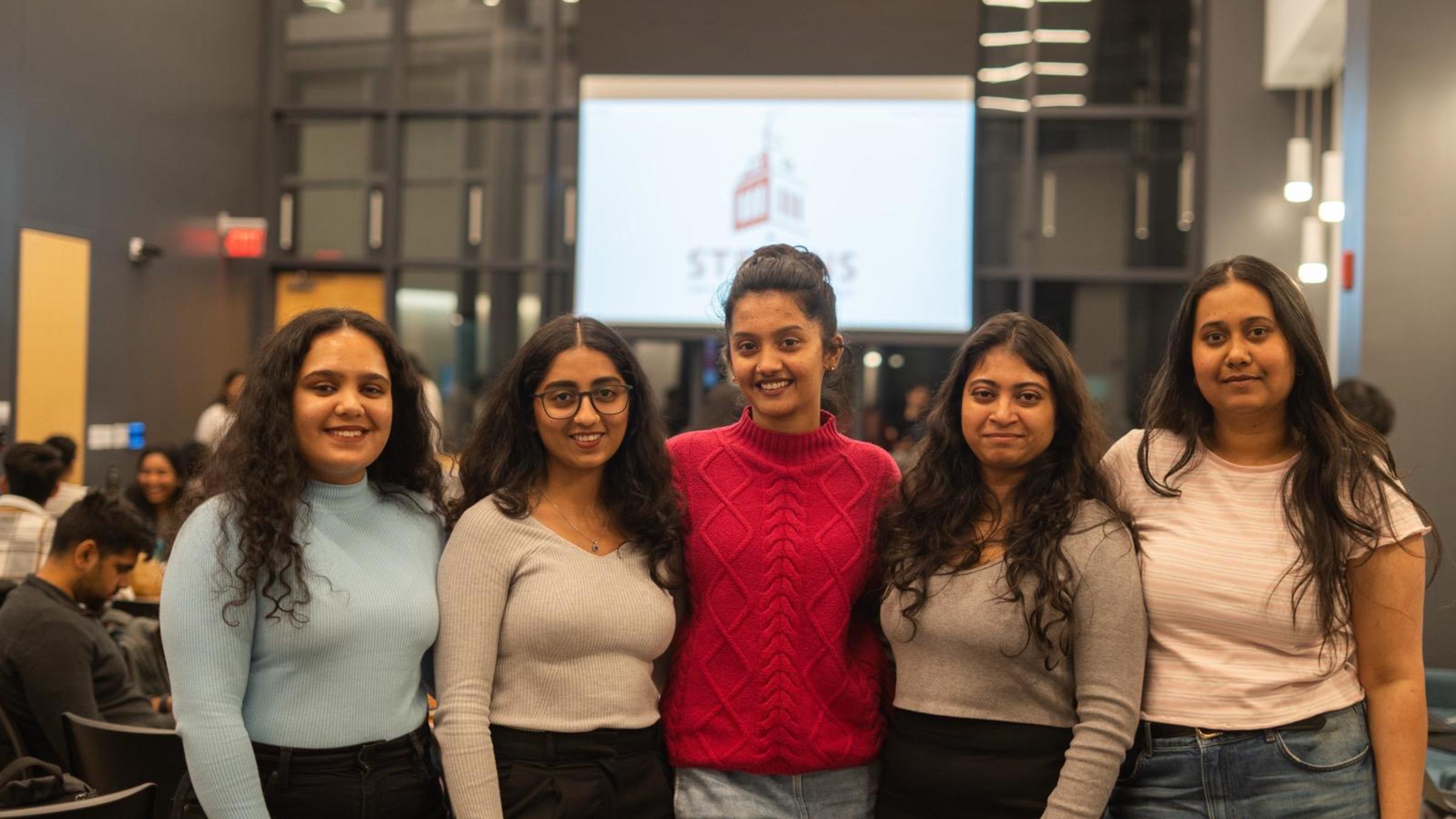 A group of five female students stand together. The camera shot is from the hips up.