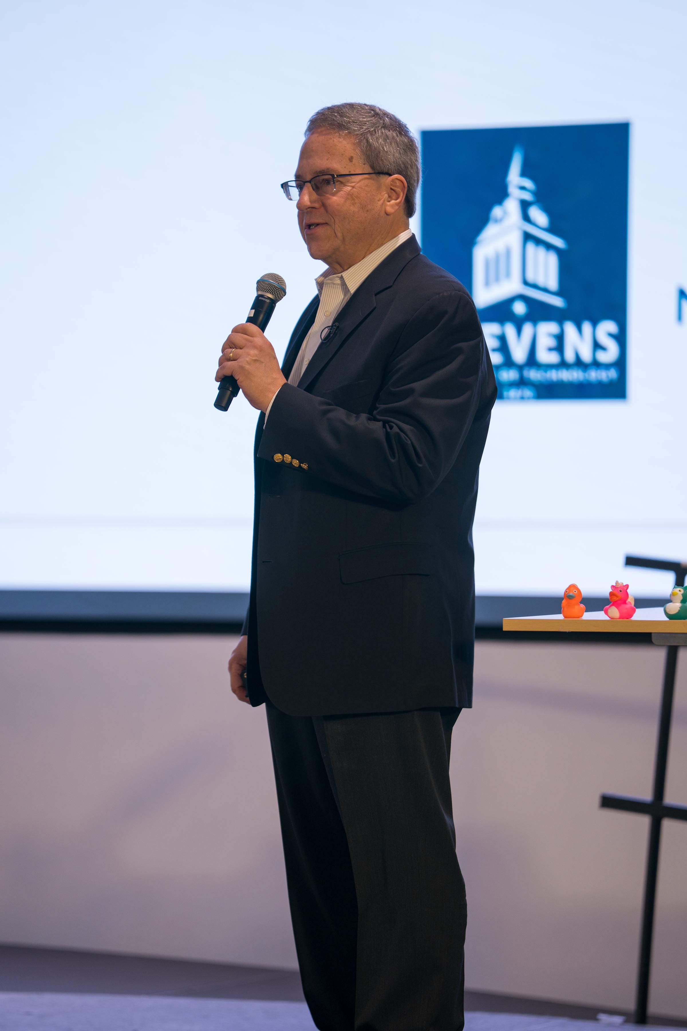 Man in black suit with microphone presents on stage, Stevens Institute logo visible on screen behind him. Colorful rubber ducks arranged on table edge at front of stage.