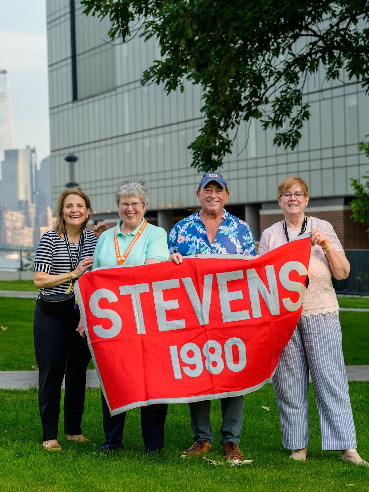 Four alumni hold a Class of 1980 banner at Alumni Weekend 2025 in front of the UCC
