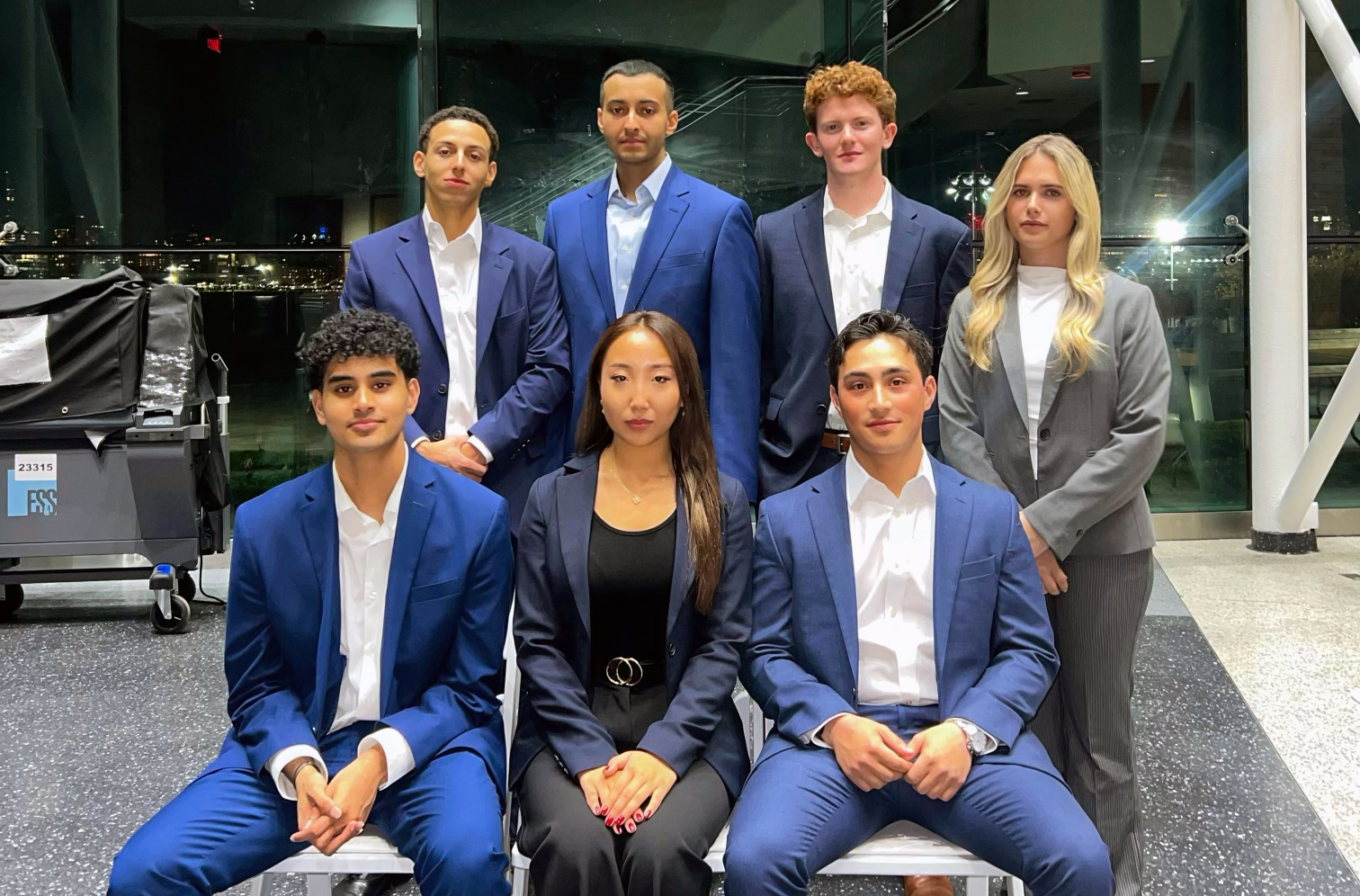 Group of seven young professionals in formal business attire posed in two rows in a modern building with glass walls at night.