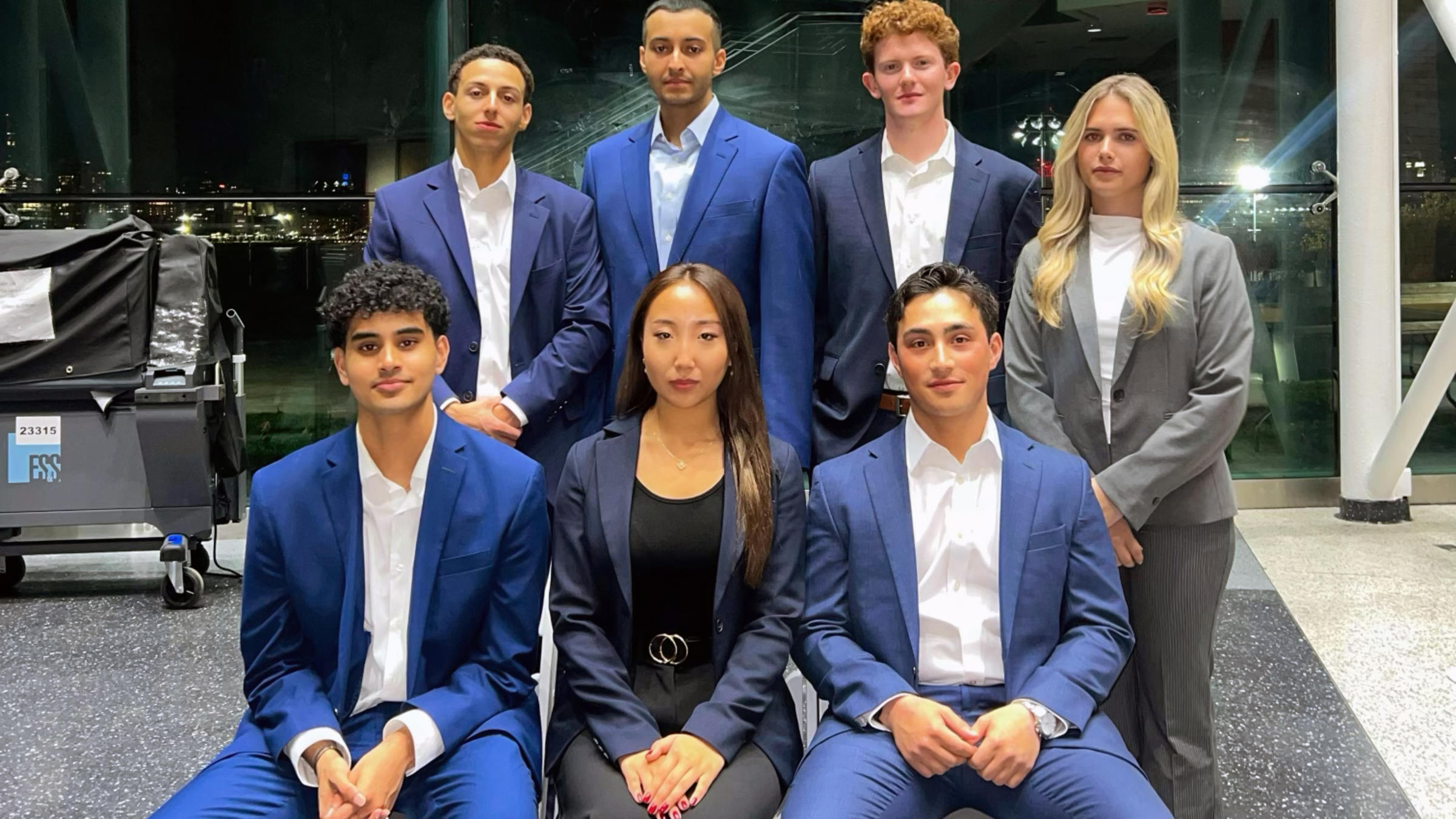 Group of seven young professionals in formal business attire posed in two rows in a modern building with glass walls at night.